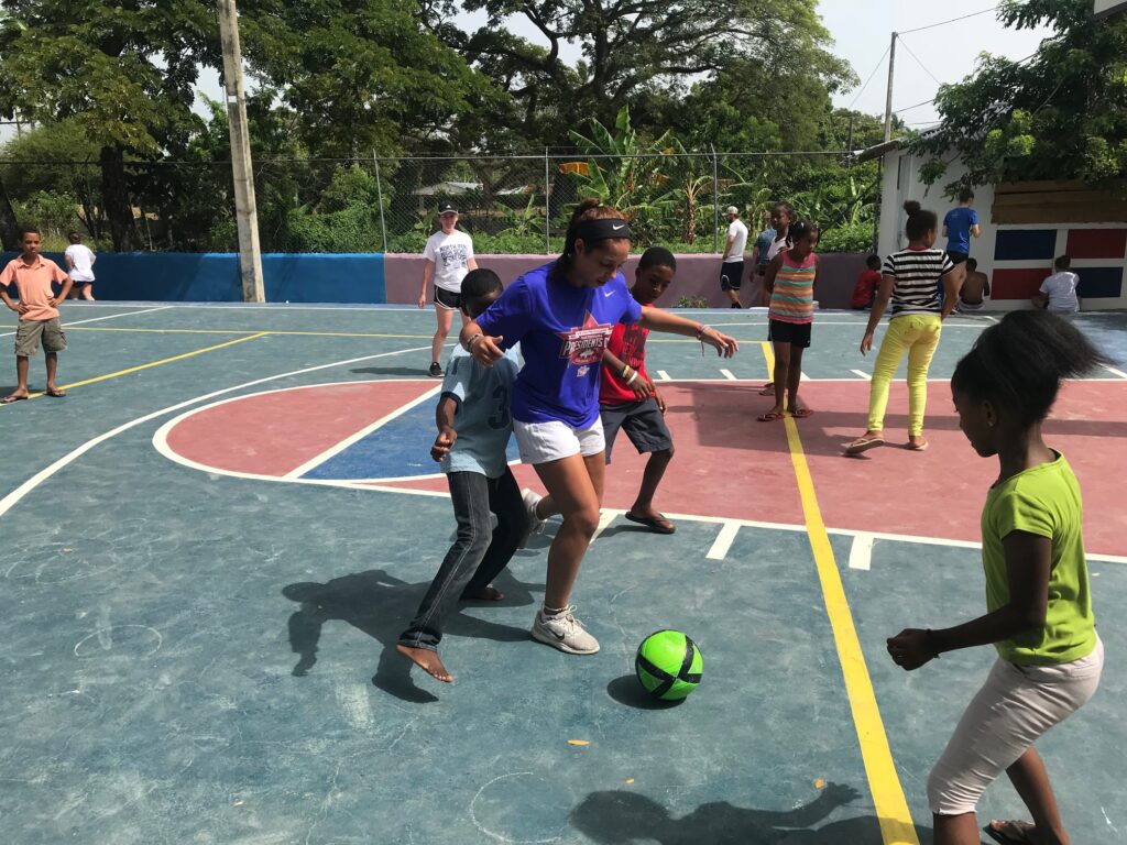 A young adult chases a green ball with children.